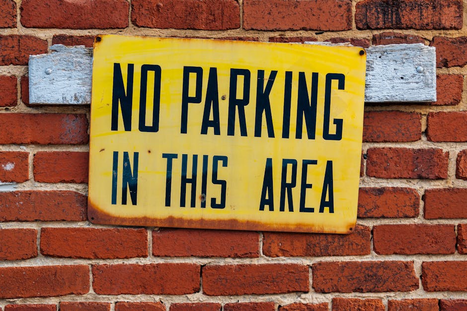 A rectangular, weathered yellow metal sign with black uppercase text reading 'NO PARKING IN THIS AREA' is mounted on a red brick wall with mortar lines. The sign is fixed to a horizontal, faded wooden plank painted light grey, which is attached to the brick surface. The background shows a typical exterior brick wall, indicating an outdoor environment, possibly near a residential or commercial property. The sign's purpose relates to parking restrictions during house removal or furniture transport, emphasizing the importance of adhering to parking regulations on moving days. Man with Van Erith, a professional house removal service, uses such signage to assist clients in planning their home relocation and avoiding parking fines, supporting efficient and compliant moving logistics involving loading areas and vehicle placement.