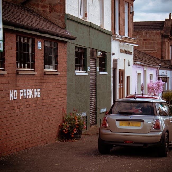 A rectangular, weathered yellow metal sign with black uppercase text reading 'NO PARKING IN THIS AREA' is mounted on a red brick wall with mortar lines. The sign is fixed to a horizontal, faded wooden plank painted light grey, which is attached to the brick surface. The background shows a typical exterior brick wall, indicating an outdoor environment, possibly near a residential or commercial property. The sign's purpose relates to parking restrictions during house removal or furniture transport, emphasizing the importance of adhering to parking regulations on moving days. Man with Van Erith, a professional house removal service, uses such signage to assist clients in planning their home relocation and avoiding parking fines, supporting efficient and compliant moving logistics involving loading areas and vehicle placement.