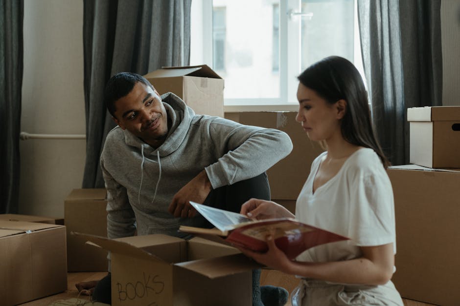 A man and a woman inside a room surrounded by stacked cardboard boxes, some labeled with handwritten notes such as 'books', indicating a home relocation or packing process. The man, wearing a grey hoo