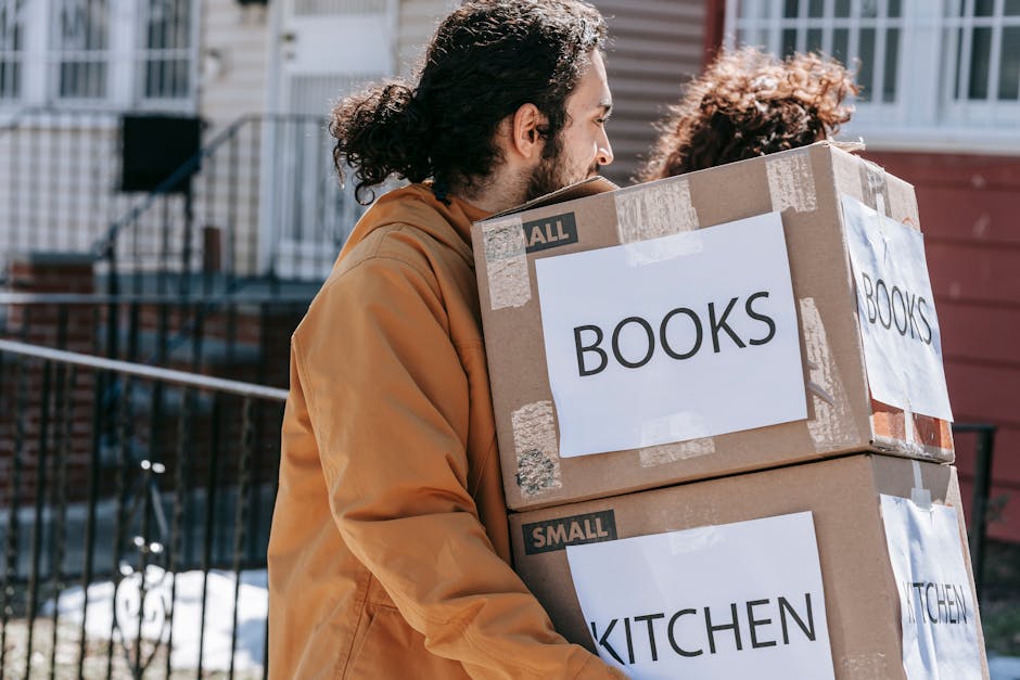 A man and a woman inside a room surrounded by stacked cardboard boxes, some labeled with handwritten notes such as 'books', indicating a home relocation or packing process. The man, wearing a grey hoo