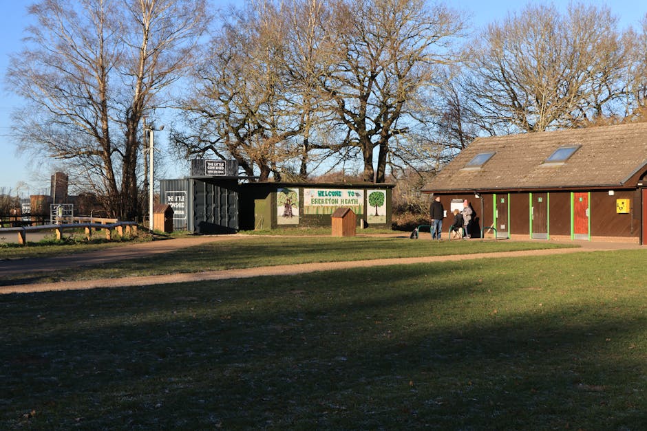A clear outdoor scene at Brereton Heath during daylight with a bright blue sky and leafless trees in the background. In the foreground, a grassy area with patches of dirt and a footpath. To the right, there is a small, brown building with green and red painted doors, along with a yellow notice board. Adjacent to the building, three people are visible; one person is handling a roller bag, while two others stand nearby, possibly engaging in a discussion or waiting. A dog is also present near the group. On the left side, there is a black, modern bus shelter with glass panels and a transparent roof, featuring signs and possibly advertising. Behind the shelter, a fence runs along the perimeter, with some industrial structures in the distance. The scene captures a moment typical of a community or park environment, with potential implications for home relocation or errands involving moving items, and features elements that could support furniture transport or packing activities, as seen in the context of house removals by Man with Van Erith.