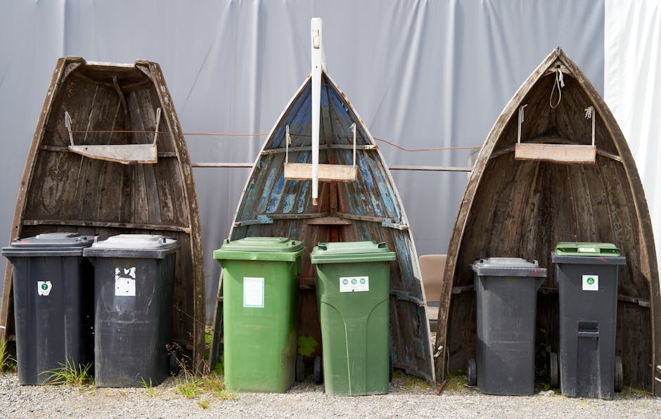 Four wooden boats, weathered and upright, positioned on a concrete surface outdoors with their bows facing upward. The boats are arranged in a row, with the central boat slightly ahead of the others, and appear to be used for decorative or storage purposes. In front of each boat, there are garbage bins—two black bins on the left and right sides, and two green bins in the center—stacked close to the boats. The green bins are marked with waste disposal symbols, indicating recycling or general waste. Behind the boats, a clothesline spans from one side to the other, and the background features a plain light-colored fabric or curtain, creating a neutral backdrop. The lighting suggests daytime, with natural light illuminating the scene. Man with Van Erith's removals service may assist with rubbish clearance or home relocation involving disposal of bulky waste or outdoor items, as indicated by the image's context.