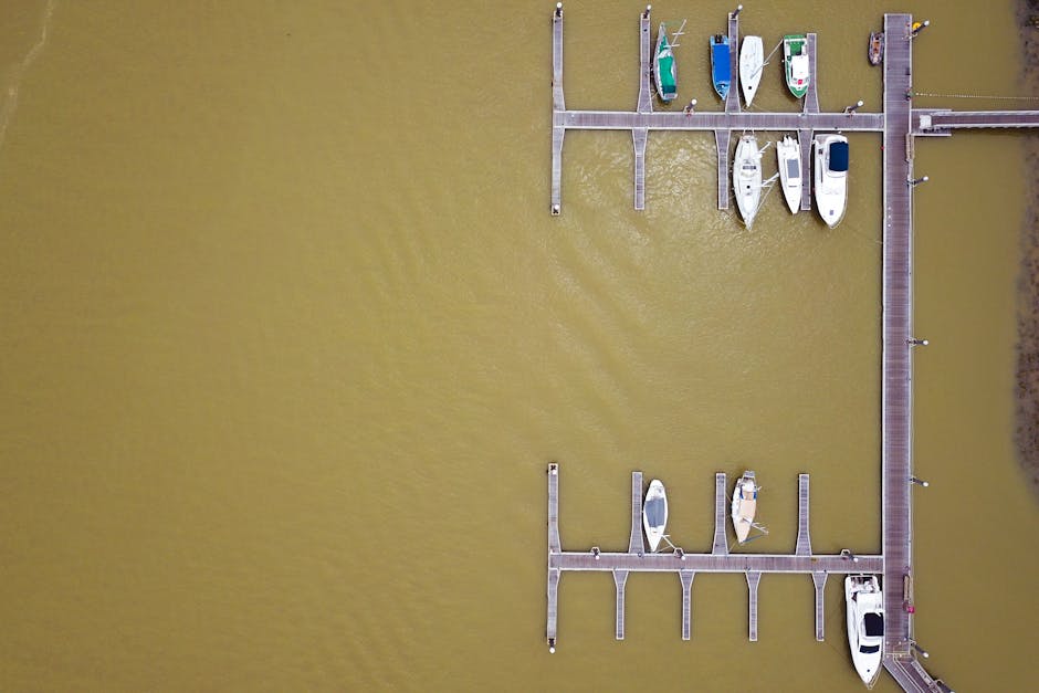 An aerial view of a marina dock with a wooden pier extending into a body of murky yellowish-brown water, where several boats of various sizes and colours, including white, blue, green, and beige, are moored at the slips. The boats are secured with ropes and are positioned parallel to the dock, with some covered by protective tarps. The surrounding landscape features a narrow strip of land with sparse vegetation on the right side of the image. The dock is equipped with small bollards and cleats for securing boats, and the wooden planks are visible along the walkway and at the boat slips. The overall scene depicts a calm, organised marina area suitable for boat storage and transportation, with evidence of recent or ongoing residential or commercial moving activities related to house removals or furniture transport, consistent with the services offered by Man with Van Erith, as seen at the provided webpage.