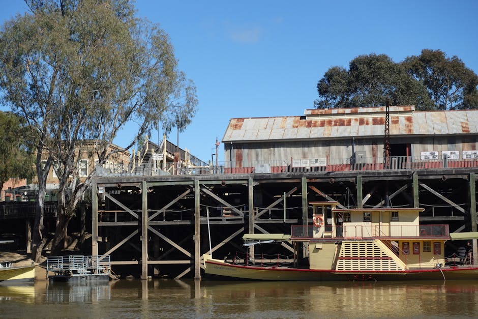 A wooden pier extending over a calm river, with weatherproofed signboards attached to a vertical wooden post near the water's edge. The signs display information related to boat fares, including prices for adults, children, under-fives, bicycles, and dogs, along with warnings about the use of the jetty and safety precautions. The pier has a handrail with additional signs such as 'Please Wait Behind This Line.' In the background, across the water, there are buildings and a small pier or dock structure, with a partly cloudy sky overhead. The scene is natural and neutral, illustrating a typical riverside location that could be part of a house removal or relocation process involving boat or ferry access, as managed by Man with Van Erith.