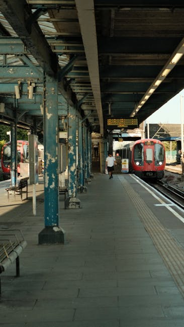 Close-up view of a railway level crossing with a red warning signal light illuminated, positioned above a brick wall with various electrical cables and conduits running along it. A metal ladder leans 