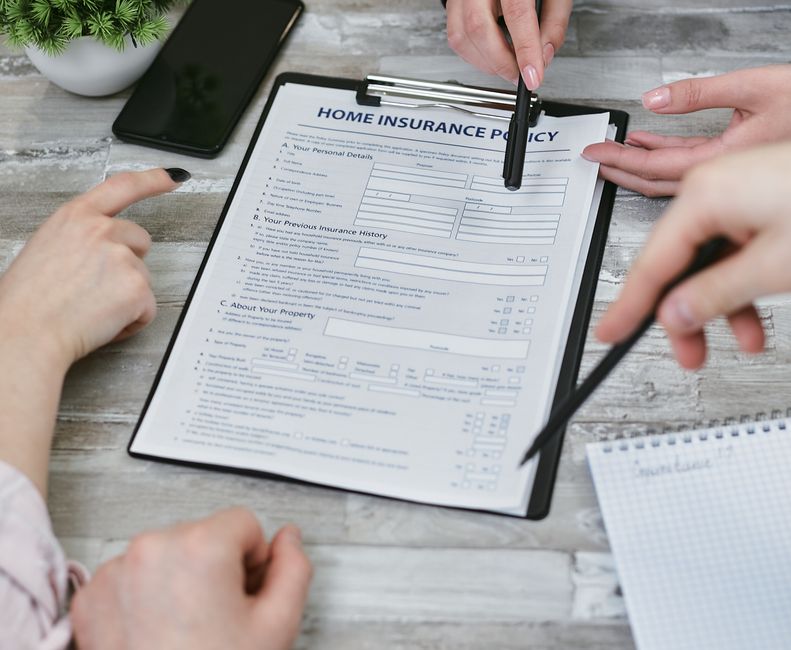 A person wearing a black jacket is seated at a light-colored wooden table, filling out a home insurance policy form secured in a black clipboard with a silver clip. They hold a black pen in their righ