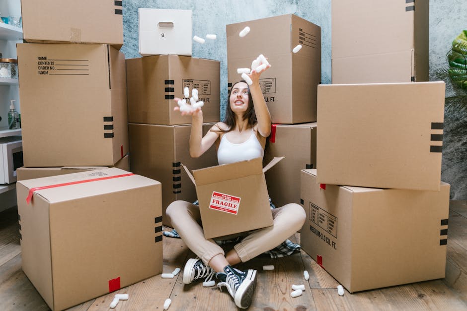 A woman sitting cross-legged on a wooden floor inside a room, surrounded by numerous cardboard moving boxes of varying sizes, some sealed with red tape and others labeled with fragile stickers, as she joyfully throws packing peanuts into the air. She is wearing a white tank top, beige trousers, and sneakers, with her head tilted back and a smile on her face. Behind her, a blue textured wall and a green potted plant are partially visible. The boxes are stacked around her, some on the floor and others placed against the wall, indicating an ongoing home relocation process supported by Man with Van Erith, specialists in removals and furniture transport, focusing on packing and moving services.