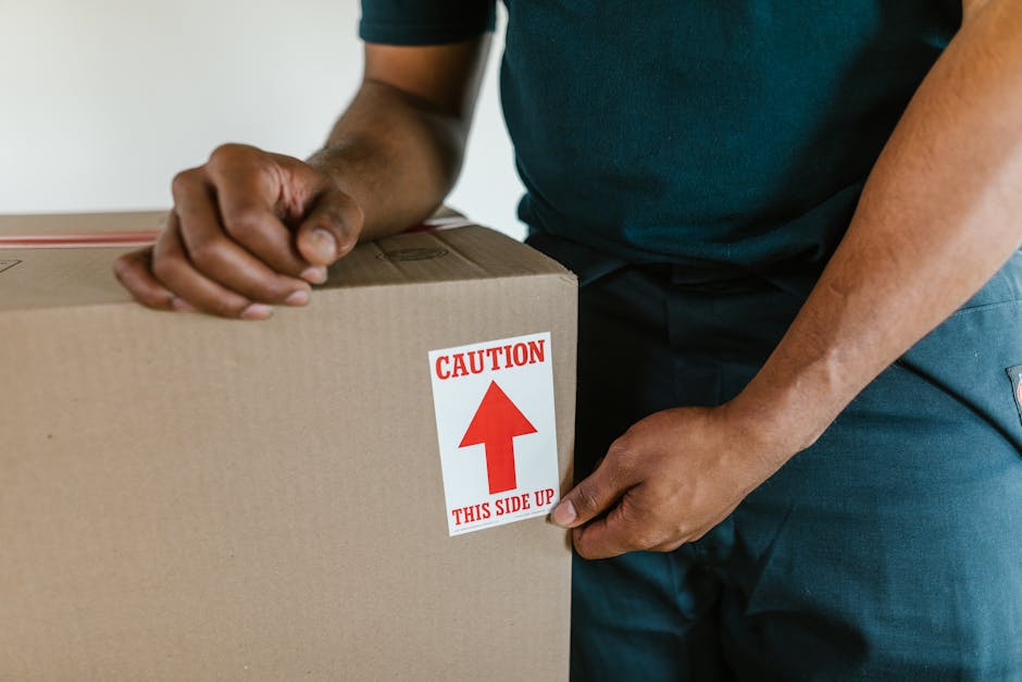 A close-up view of a person wearing a dark green t-shirt and blue work trousers, holding a large cardboard box during a home relocation process. The person is gripping the box with both hands while positioning it for transportation, with the left hand on the side and the right hand supporting the top. The cardboard box has a red and white caution label prominently displayed on the front, indicating 'Caution' with an upward arrow and the instruction 'This side up.' Inside the property, the background shows a partially visible interior space with neutral-colored walls, suggesting that the scene takes place during packing or furniture moving. The box appears sturdy and is packed for moving, possibly containing fragile or valuable items, aligned with furniture transport and packing procedures carried out by [COMPANY_NAME]. The overall environment is well-lit, emphasizing careful handling and professional loading activities typical of house removals and moving services.