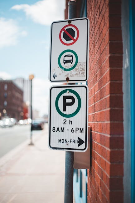 A street scene featuring a vertical metal signpost mounted on a brick wall, displaying parking restrictions and parking allowable times. The upper part of the sign has two additional rectangular panels: one with a no-parking symbol crossed out in red, indicating a parking restriction or prohibition, and another with a bus icon within a circle, denoting bus parking or stopping rules. The lower panel shows a white background with a large green letter 'P' and text specifying parking is permitted for up to 2 hours between 8am and 6pm from Monday to Friday. The background reveals a city street with blurred cars, buildings, and a partly cloudy sky, suggesting an urban environment suitable for house removals or furniture transport, where adherence to parking regulations is essential for smooth loading and unloading during home relocation services by Man with Van Erith.