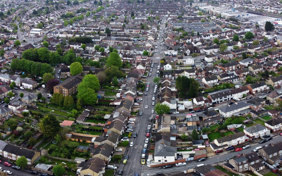 An aerial view of a residential neighbourhood with densely packed houses, trees, and greenery, showing a straight street lined with parked cars on both sides. The street runs from the foreground to the background, with houses featuring pitched roofs, gardens, and driveways. Some properties have visible fencing, outdoor furniture, and small sheds. The area is illuminated by daylight, with natural lighting highlighting the rooftops and foliage. This scene depicts a typical home environment suitable for property relocations, supported by local infrastructure and community layout, relevant to house removals and moving services such as those provided by Man with Van Erith.