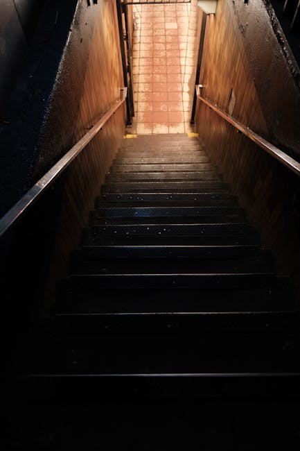 A narrow staircase inside a property, viewed from the top looking down, with wooden handrails mounted on each side and dark, possibly rubber or carpeted steps showing signs of wear. The staircase leads to a brightly lit area with terracotta floor tiles, suggesting an entrance or exit area. Adjacent to the staircase, the walls are lined with wooden panels, and there is a dark surrounding space indicating the staircase is enclosed. The environment is lit by natural or warm artificial light, highlighting the textures of the wooden handrails and the steps. This scene captures a typical home relocation or furniture transport setting, where careful handling of household items through tight spaces is necessary, consistent with the services offered by Man with Van Erith for removals and moving logistics.