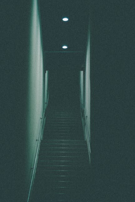 A narrow staircase inside a residential property, featuring dark wooden steps and light-colored walls on either side, illuminated by three small ceiling-mounted lights. The staircase appears to be part of a home in an urban setting, likely within a flat or apartment. At the base of the stairs, a moving trolley loaded with cardboard boxes and wrapped furniture is positioned, indicating an active home relocation or packing process. The boxes are sealed with packing tape, and some are stacked on top of each other, ready for transport by a professional moving company. The scene suggests careful handling and preparation for furniture transport in a confined space, typical of urban house removals. The environment appears clean and secure, with no clutter visible around the stairs, aligning with a professional move conducted by Man with Van Erith for a client in Erith. The lighting and composition highlight the logistics involved in navigating tight spaces during a house move, emphasizing the importance of skilled furniture transport and packing services.