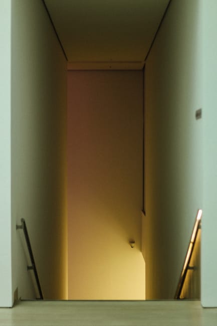 A narrow staircase inside a property, viewed from the top looking down, with wooden handrails mounted on each side and dark, possibly rubber or carpeted steps showing signs of wear. The staircase leads to a brightly lit area with terracotta floor tiles, suggesting an entrance or exit area. Adjacent to the staircase, the walls are lined with wooden panels, and there is a dark surrounding space indicating the staircase is enclosed. The environment is lit by natural or warm artificial light, highlighting the textures of the wooden handrails and the steps. This scene captures a typical home relocation or furniture transport setting, where careful handling of household items through tight spaces is necessary, consistent with the services offered by Man with Van Erith for removals and moving logistics.